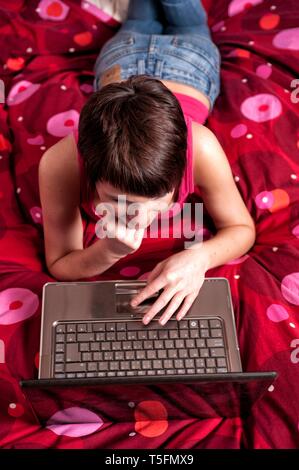 Junge Frau liegt auf ihrem Bett mit einem Leptop vor sich vor sich und schaut in die freundlich in die Kamera.
