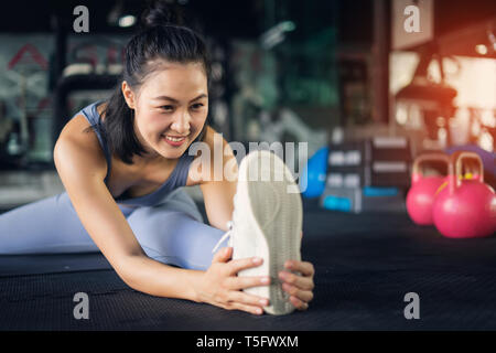 Young asian woman in gym healthy lifestyle sitting on yoga mat holding foot stretching leg. exercise and health concept in fitness gym.