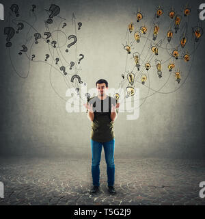 Perplexed young man has different questions and ideas as holding interrogation marks in one palm and light bulbs in another. Unsure thoughtful guy mak Stock Photo