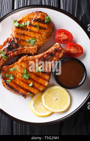 Delicious grilled pork steak in a spicy tamarind sauce, with lemon and fresh tomatoes closeup on a plate on the table. Vertical top view from above Stock Photo