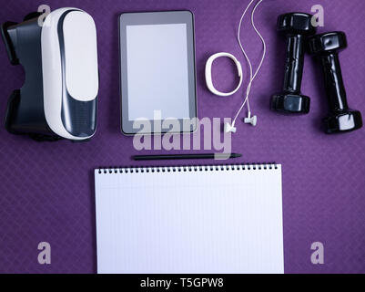 Closeup on VR glasses, white fitness tracker, headphones, tablet PC, black dumbbells and opened notebook with black pen laying on purple fitness mat.  Stock Photo