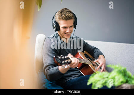 Smiling young man with headphones sitting on couch playing guitar Stock Photo