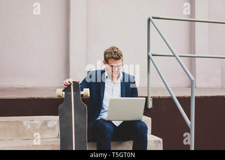 Smiling young businessman with skateboard sitting outdoors on stairs using laptop Stock Photo