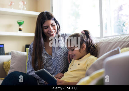 Happy mother and daughter sitting on couch at home using tablet Stock Photo