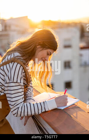 Smiling young woman taking notes on roof terrace at sunset Stock Photo