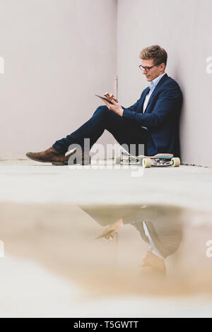 Smiling young businessman with skateboard sitting outdoors using tablet Stock Photo