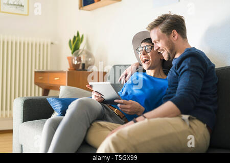 Happy casual couple relaxing on couch using tablet and laughing Stock Photo