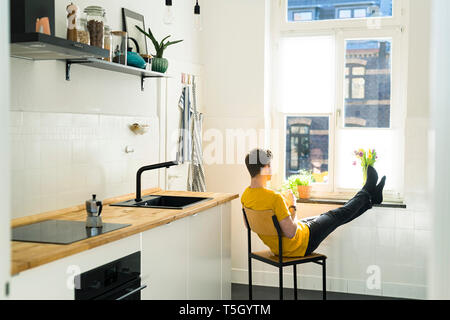 Man relaxing on chair with his feet on the windowsill in his kitchen looking outside Stock Photo