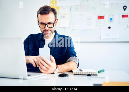 Casual businessman using smartphone and laptop at desk in office Stock Photo