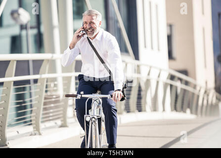 Happy mature businessman talking on cell phone and riding bicycle on a bridge in the city Stock Photo