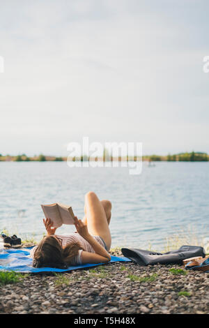 Young woman lying on blanket at lakeshore reading a book Stock Photo