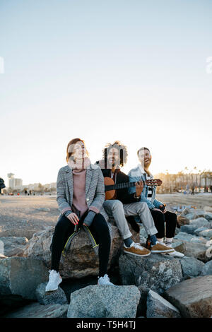 Three happy friends with guitar sitting outdoors at sunset Stock Photo