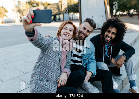 Three happy friends sitting outdoors taking a selfie Stock Photo