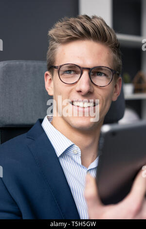 Smiling young businessman sitting on office chair using tablet Stock Photo