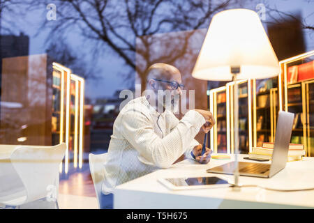 Mature man sitting at desk in a library in the evening using laptop Stock Photo