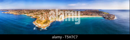 Famous Bondi beach on Sydney Pacific coast in elevated aerial wide panorama between sandstone headlands with distant city CBD on the horizon.
