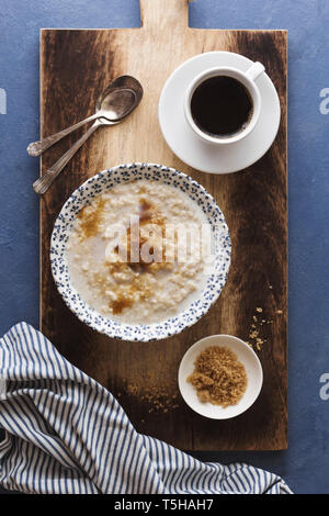 A board and napkin with a bowl of oatmeal porridge and a cup of coffee, two spoons, and brown sugar Stock Photo