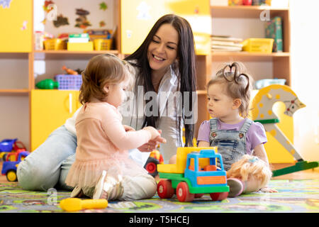 Children kids toddlers playing with teacher in kindergarten. Nursery babies sitting on floor together with caregiver Stock Photo