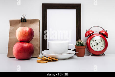 Mock Up dark photo frame, red alarm clock, paper bag, two apples, crackers and a white cup of coffee with a saucer on a light background. Stock Photo