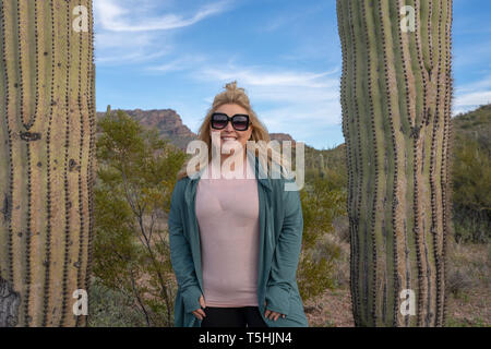 Happy, relaxed blond woman wearing sunglasses stands between two saguaro cactus (cacti) in Arizona, wearing casual clothing Stock Photo