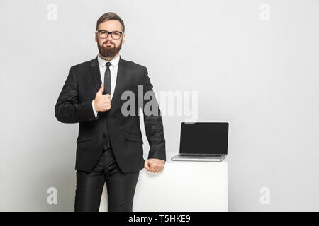 Portrait of handsome confident successful bearded young businessman in intelligence black suit are standing near his working place and showing thumbs  Stock Photo