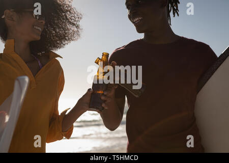 Couple toasting beer bottle while holding skateboard at beach Stock Photo