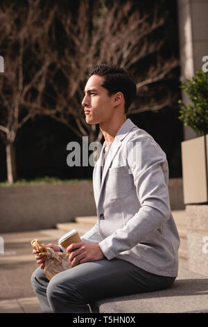 Young man having coffee and breakfast while sitting on stone bench Stock Photo