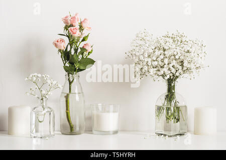 Composition with white and pink flowers in glass bottles and candles on table wall background. Stock Photo