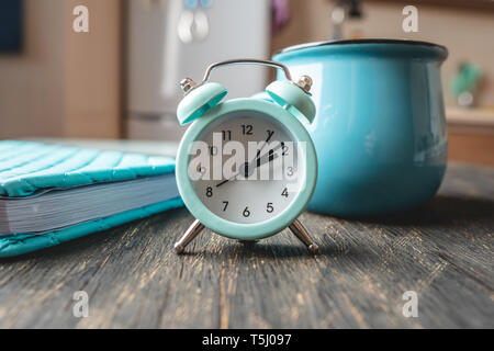 Blue metal alarm clock on the table with a mug and a diary on the background of the kitchen. Concept of time and home mode Stock Photo