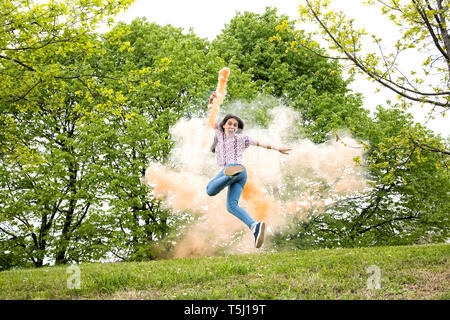 Agile excited young girl with a colored smoke flare running and jumping for joy in a wooded park or garden Stock Photo