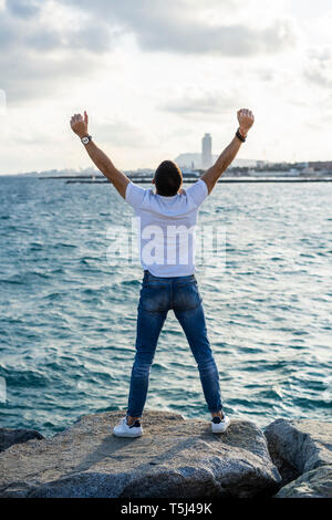 Spain, Barcelona, back view of man with raised arms standing in front of the sea Stock Photo