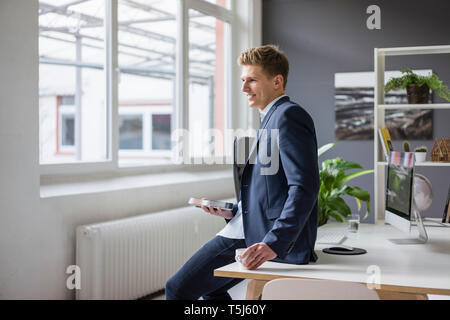 Smiling young businessman sitting on desk in office having a coffee break Stock Photo