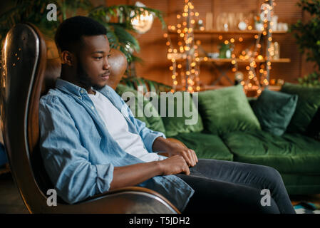 Black man sitting in a comfortable leather chair in the living room, relaxation at home. Young african american male person in his house Stock Photo