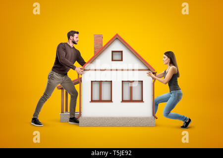 Young man and woman in casual clothes holding a big white house with red roof on yellow background Stock Photo