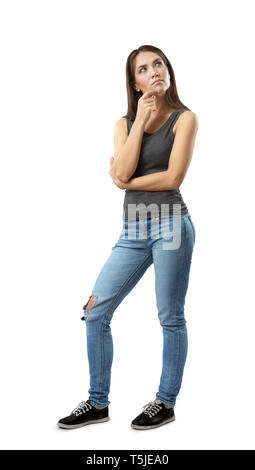Woman in gray top and blue jeans standing with thoughtful expression on her face and with hand on her chin looking up isolated on white background. Stock Photo