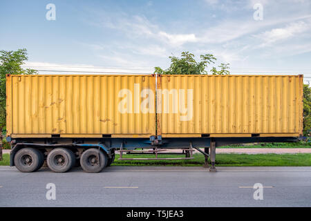 A metal trailer is parked on a road next to a white house and green ...