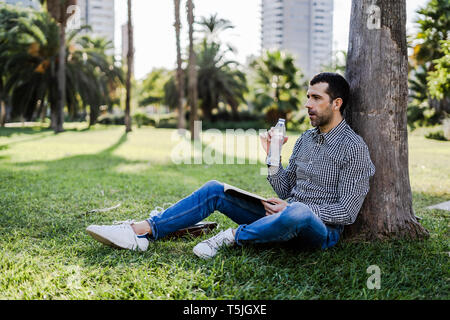 Man on a meadow in city park leaning against tree drinking water Stock Photo