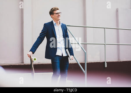 Smiling young businessman with skateboard and laptop standing outdoors on stairs Stock Photo