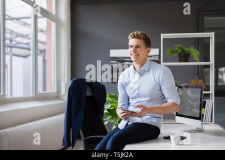 Smiling young businessman sitting on desk in office holding tablet Stock Photo