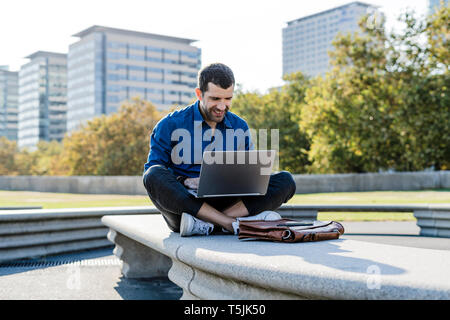 Smiling businessman sitting on bench outdoors working on laptop Stock Photo