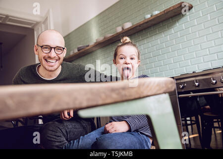 Father and daughter sitting in kitchen, girl pulling funny faces Stock Photo
