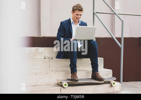 Happy young businessman with skateboard sitting outdoors on stairs using laptop Stock Photo
