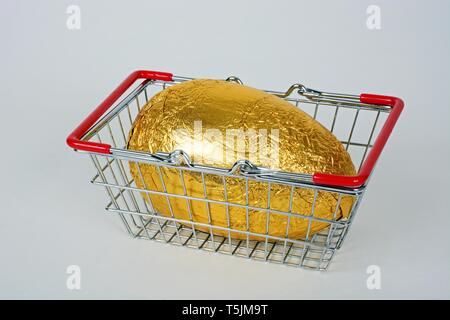 Chocolate Easter egg wrapped in gold foil in a shopping basket against a white background, UK. Stock Photo
