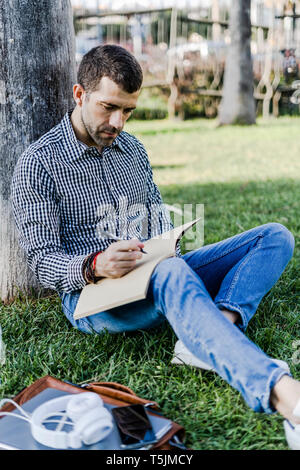 Man on a meadow leaning against tree trunk taking notes Stock Photo