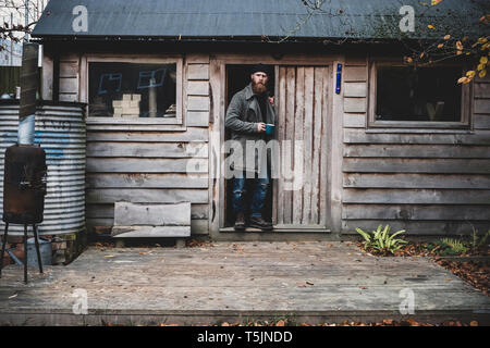 Bearded man standing in doorway of wooden workshop, holding blue mug, looking at camera. Stock Photo