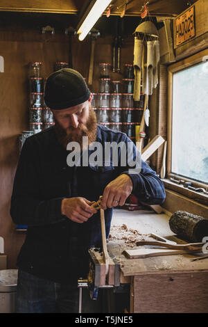 Bearded man wearing black beanie standing at workbench in workshop, working on piece of wood. Stock Photo