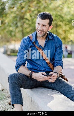 Portrait of smiling man with smartphone sitting on a wall Stock Photo