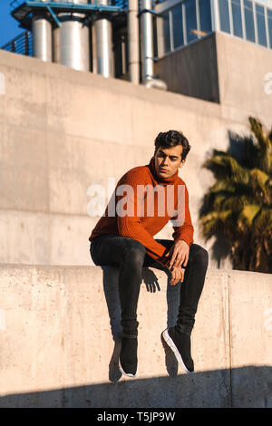 Portrait of fashionable young man wearing turtleneck pullover sitting on a wall at sunlight Stock Photo