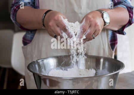 Close up of person wearing apron standing in kitchen, mixing ingredients for a crumble in metal bowl. Stock Photo