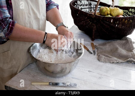 Close up of person wearing apron standing in kitchen, mixing ingredients for a crumble in metal bowl. Stock Photo